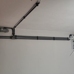Krzysztof Wilk Elektryka i Automatyka Domowa - Czarna instalacja kablowa na białym suficie i ścianie, z szarą rurą i białą skrzynką z kablami. Widok z dołu, ukazujący szczegóły montażu.