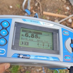 Krzysztof Wilk Elektryka i Automatyka Domowa - Miernik Metrel MI 3102 BT EurotestXE pokazuje wynik pomiaru uziemienia 6.85 Ω. Widoczne kable i fragment podłoża. Urządzenie w dłoni.