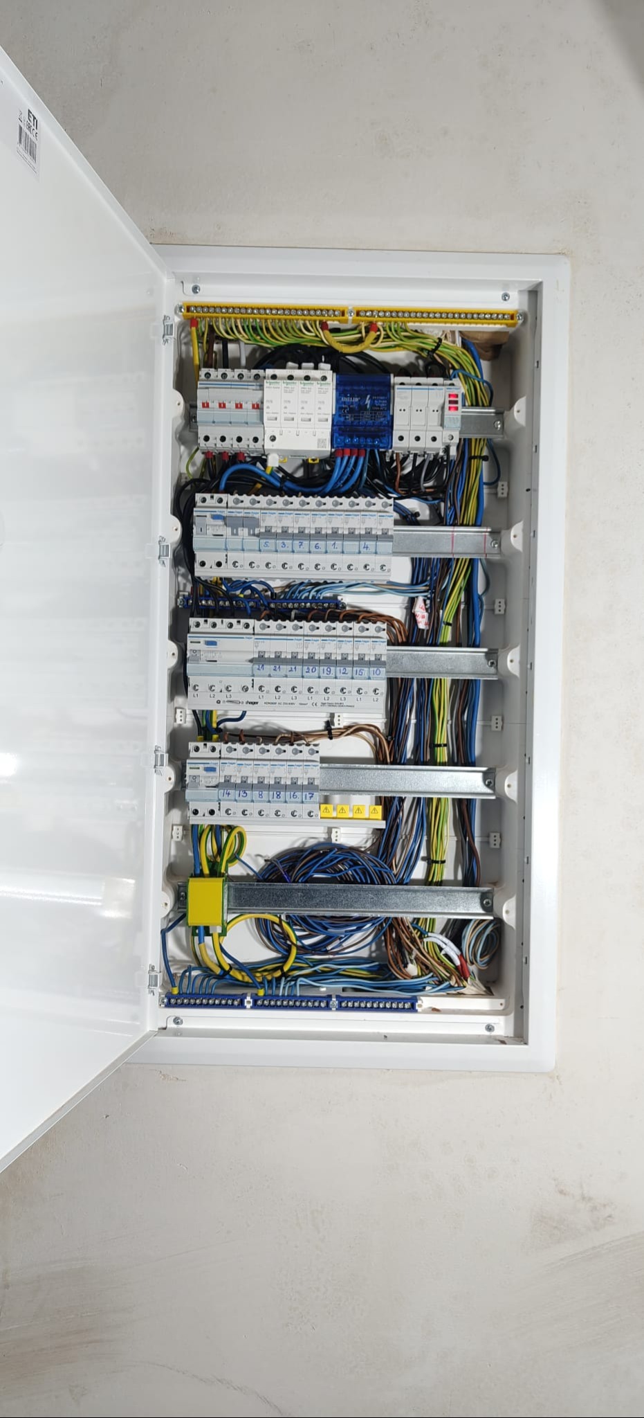 Otwarta, biała skrzynka rozdzielcza z widocznymi bezpiecznikami, przewodami w kolorach niebieskim, żółtym i zielonym. Widok z bliska na instalację elektryczną.