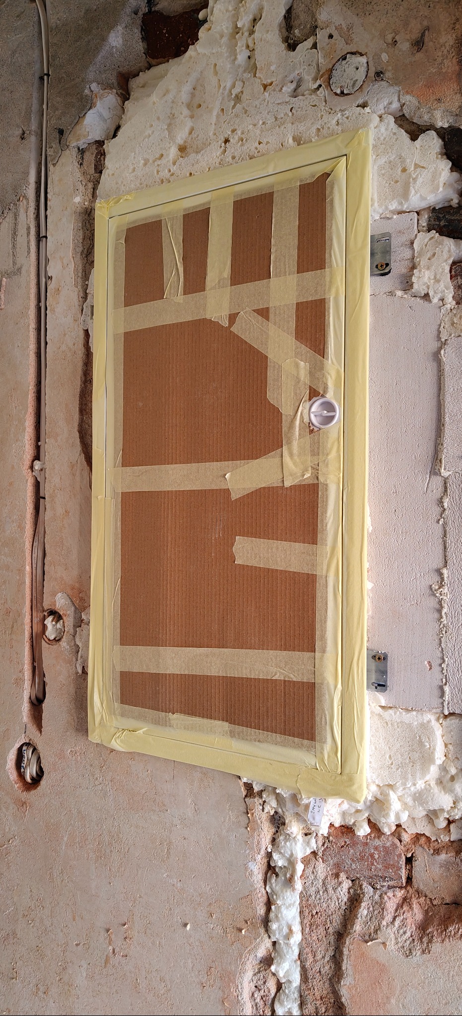 Zabezpieczona klapa rewizyjna na ścianie z widocznymi przewodami elektrycznymi i pianą montażową. Prace wykończeniowe w trakcie realizacji.