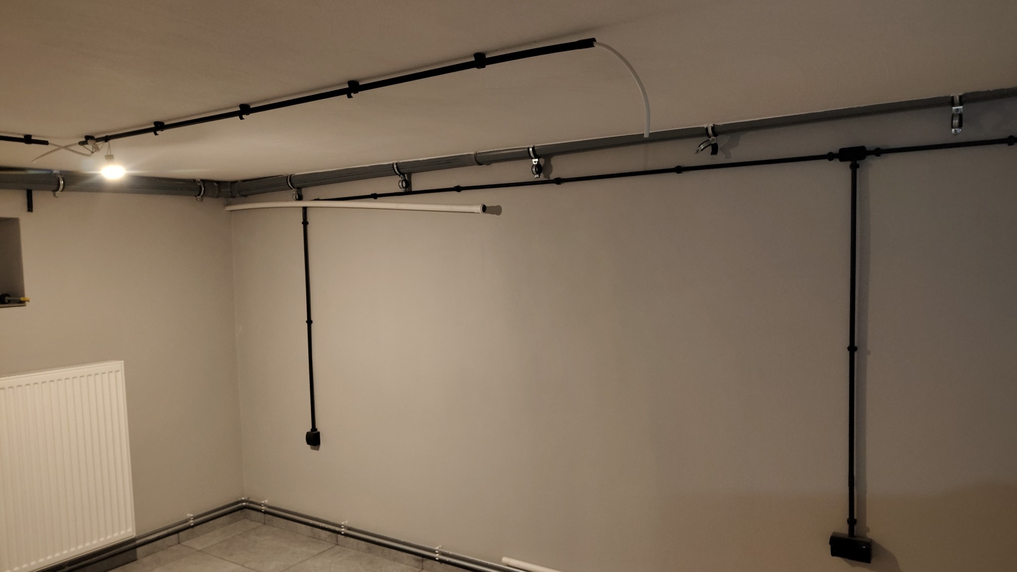 Szara ściana z widoczną instalacją elektryczną w czarnych rurkach, poprowadzona po suficie i ścianach. Widoczny fragment grzejnika i podłogi z szarych płytek.