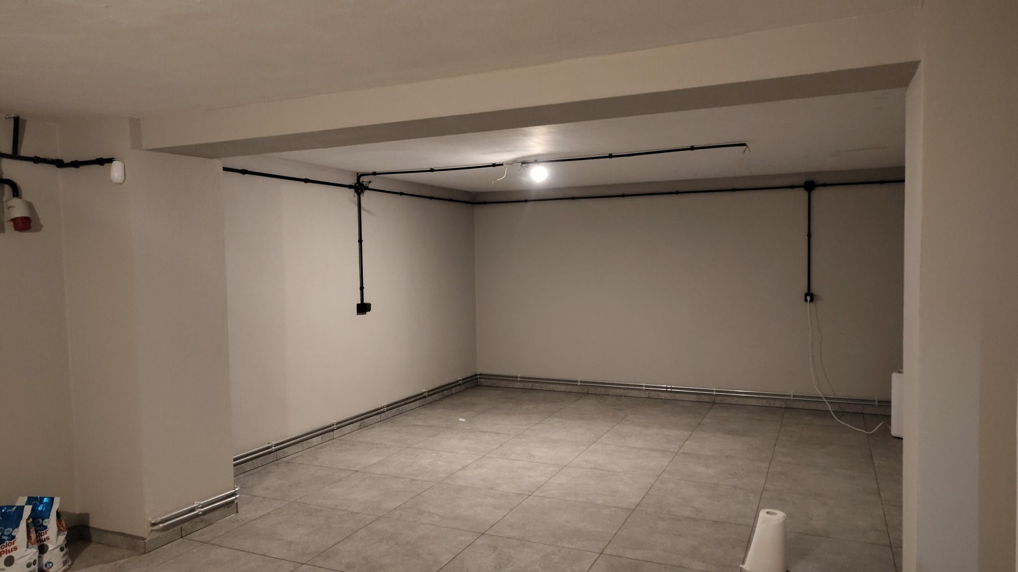 Wykończone wnętrze z widoczną, natynkową instalacją elektryczną w czarnych rurkach. Na ścianach gniazdka i włączniki. Pod sufitem lampa. W tle widoczne worki z zaprawą.