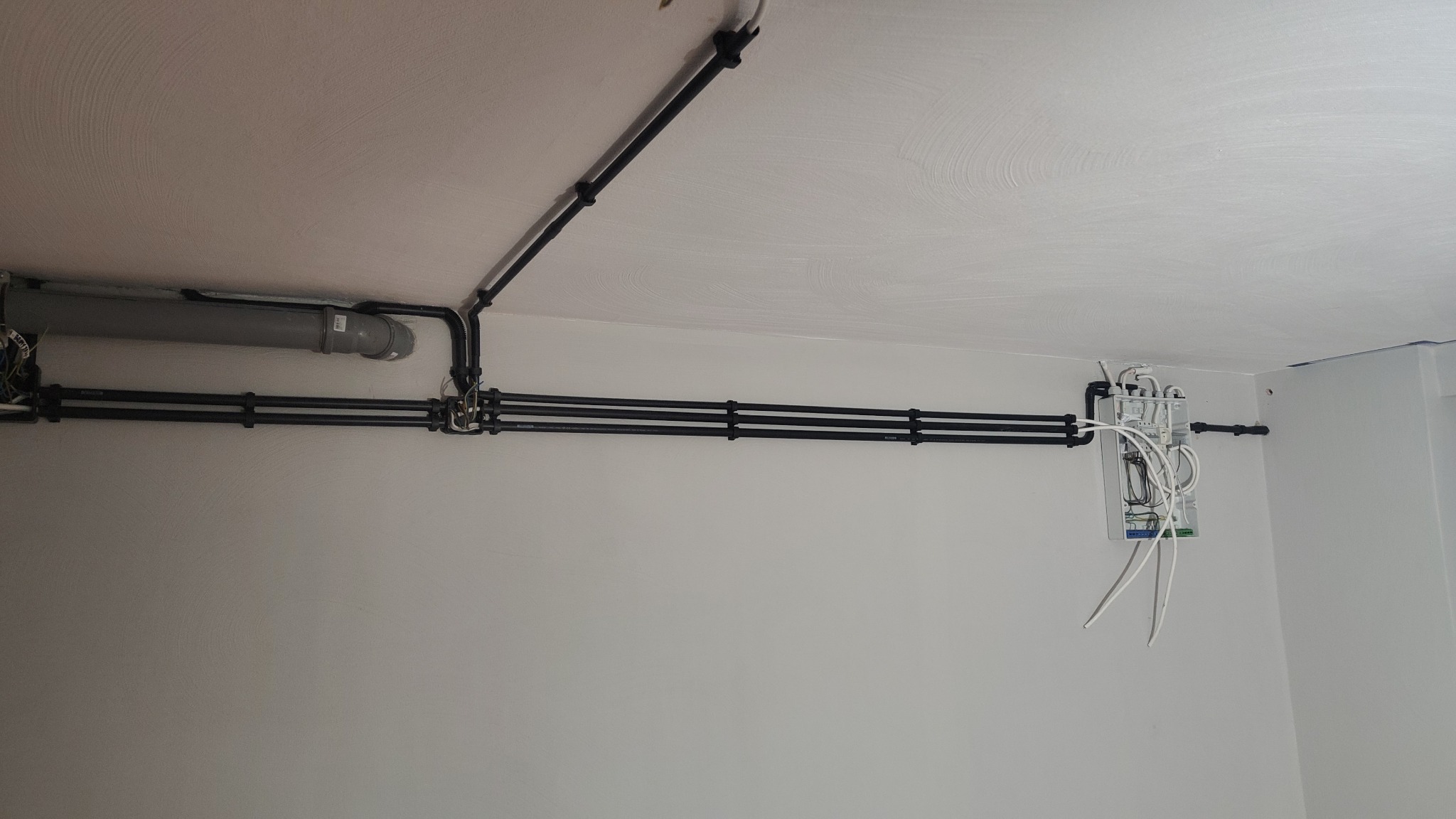 Czarna instalacja kablowa na białym suficie i ścianie, z szarą rurą i białą skrzynką z kablami. Widok z dołu, ukazujący szczegóły montażu.