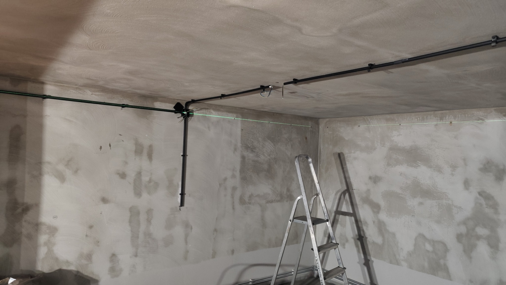 Montaż instalacji elektrycznej w surowym wnętrzu. Widoczne rury, laser krzyżowy wyznaczający poziom, drabina. Ściany i sufit w stanie deweloperskim.