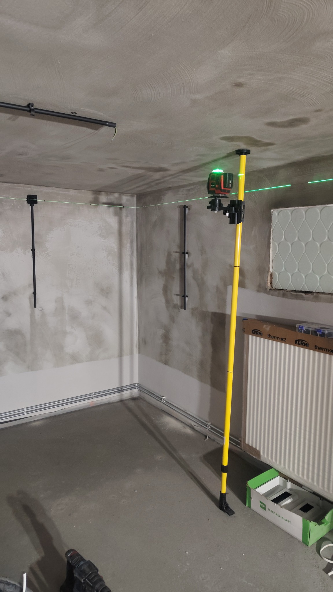Wnętrze w trakcie prac wykończeniowych z widocznym laserem krzyżowym na żółtym statywie, wyznaczającym poziom. Na ścianach rury i uchwyty instalacyjne.