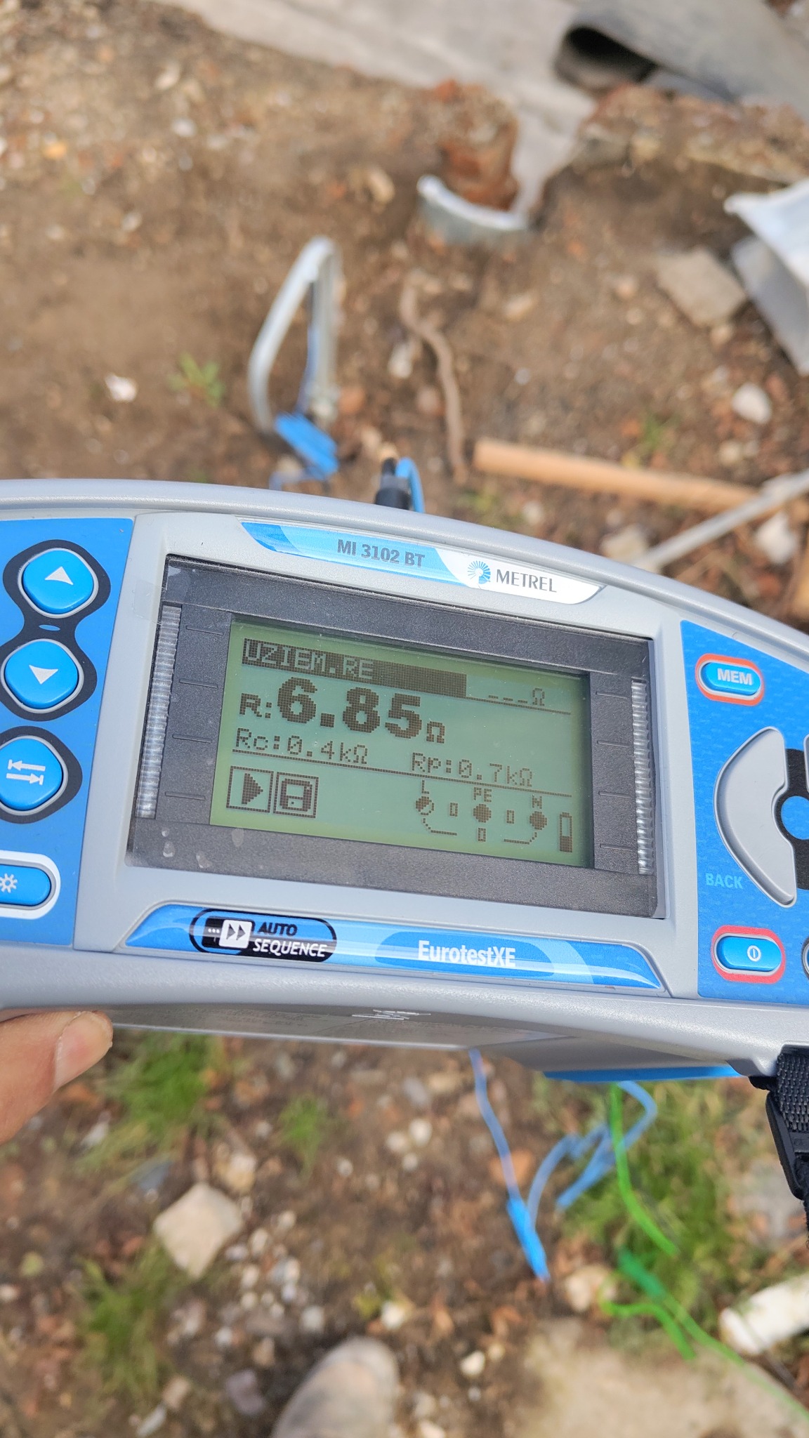 Miernik Metrel MI 3102 BT EurotestXE pokazuje wynik pomiaru uziemienia 6.85 Ω. Widoczne kable i fragment podłoża. Urządzenie w dłoni.