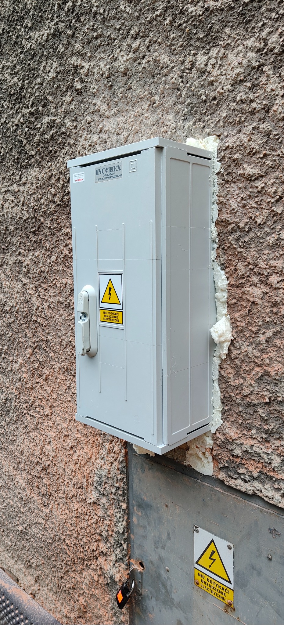 Szara skrzynka elektryczna INCOBEX zamontowana na chropowatej ścianie, uszczelniona pianką montażową. Widoczny zamek i ostrzeżenie o urządzeniu elektrycznym.