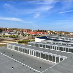 Restormann Zakrzewski Sp.k - Płaski dach budynku przemysłowego z świetlikami, widok z góry na tle zabudowy miejskiej i błękitnego nieba. Dach pokryty papą, z widocznymi elementami mocującymi.