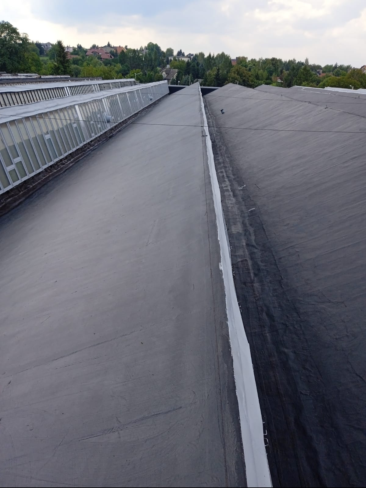 Dach przemysłowy z widoczną membraną bitumiczną i fragmentem dachu szklanego. Widoczne łączenia i detale konstrukcyjne. Tło: zabudowa i zieleń.