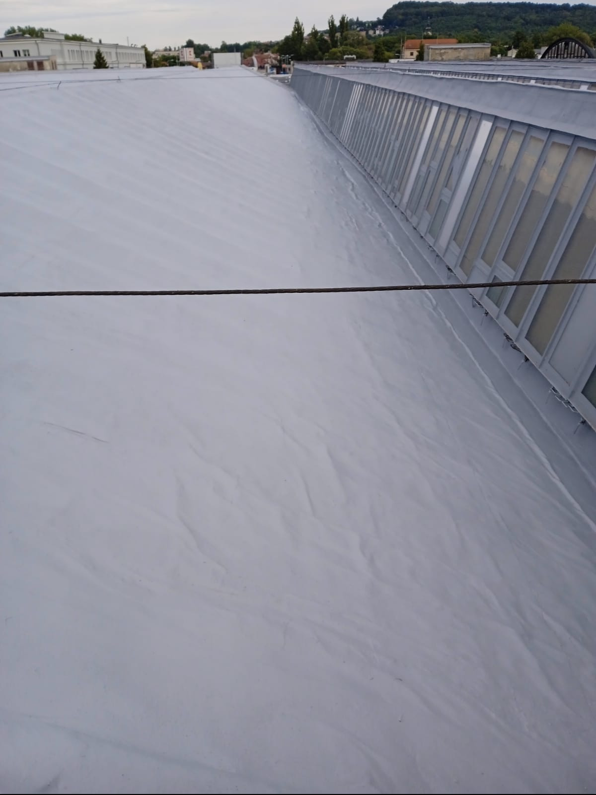 Szary, płaski dach przemysłowy z widocznymi oknami dachowymi i pojedynczym przewodem biegnącym w poprzek. Widok z góry na pokrycie dachowe.