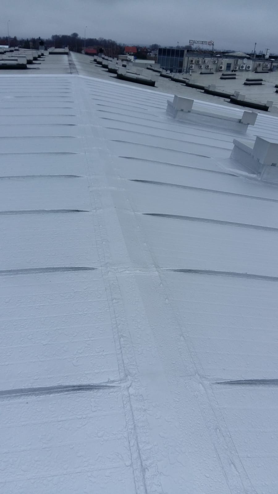 Szary, metalowy dach przemysłowy z widocznymi łączeniami i elementami wentylacyjnymi, sfotografowany z góry w pochmurny dzień. Widoczny budynek w tle.