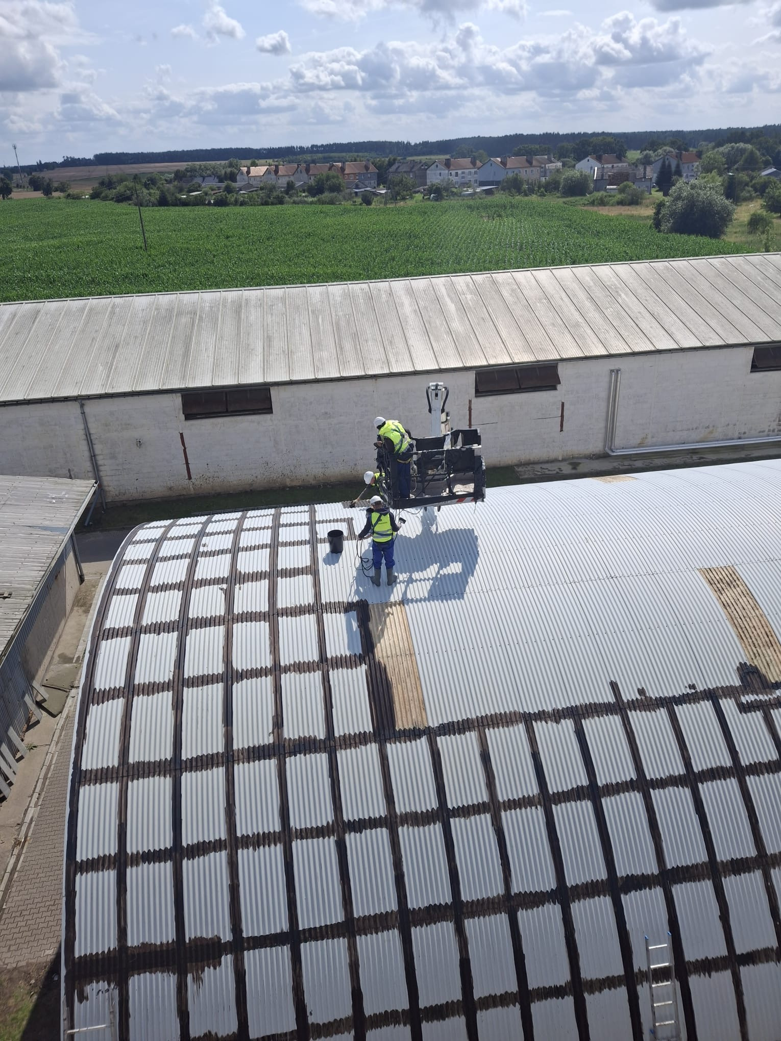 Prace na dachu z blachy falistej: dwóch robotników w żółtych kamizelkach nanosi czarną substancję na łączenia blach z podnośnika koszowego. W tle budynki gospodarcze i pola.