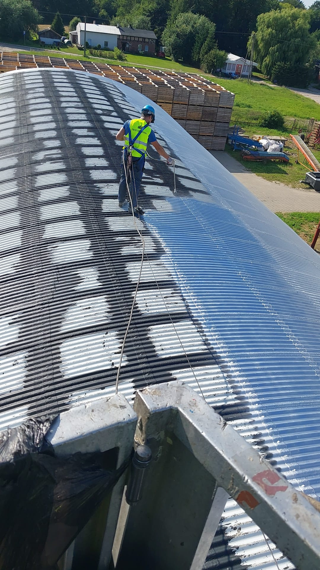 Pracownik w uprzęży maluje metalowy dach magazynu z wyraźnymi śladami korozji. Częściowo pomalowana powierzchnia kontrastuje z niepomalowaną.