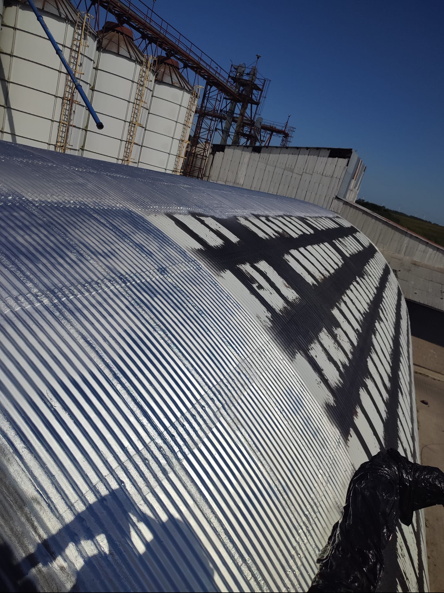 Metalowy dach przemysłowy po renowacji w Gdyni. Widoczne ślady świeżo nałożonej powłoki ochronnej i elementy konstrukcji silosów w tle. Ujęcie z góry.