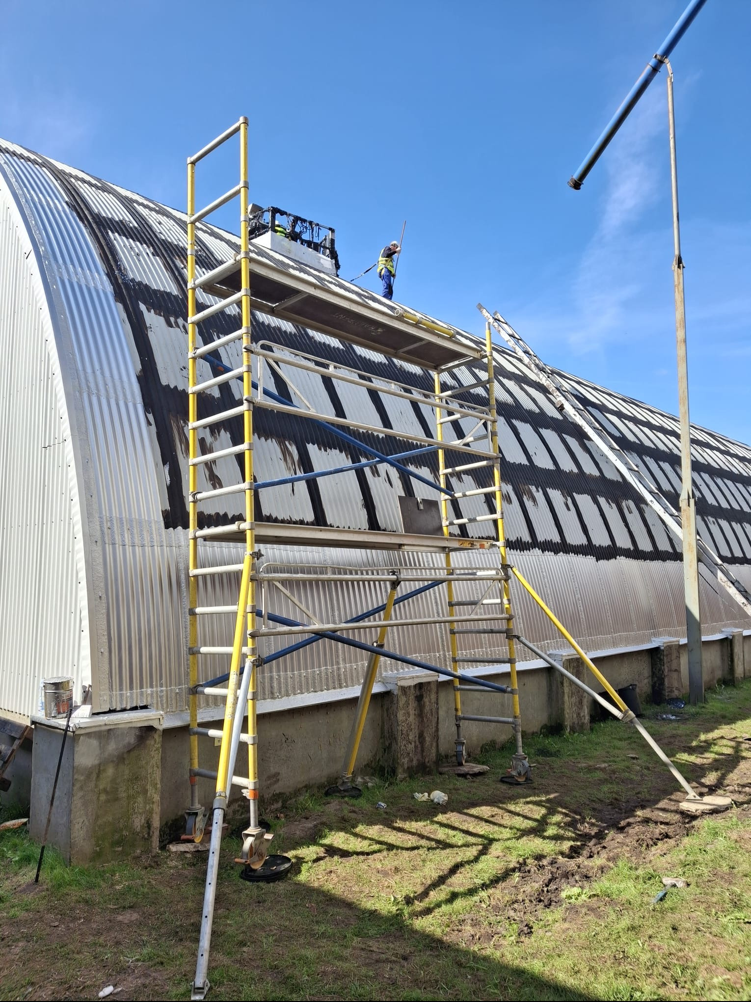 Pracownik na rusztowaniu maluje dach z blachy falistej. Widoczny fragment pomalowanej powierzchni i narzędzia pracy. Niebo bezchmurne.