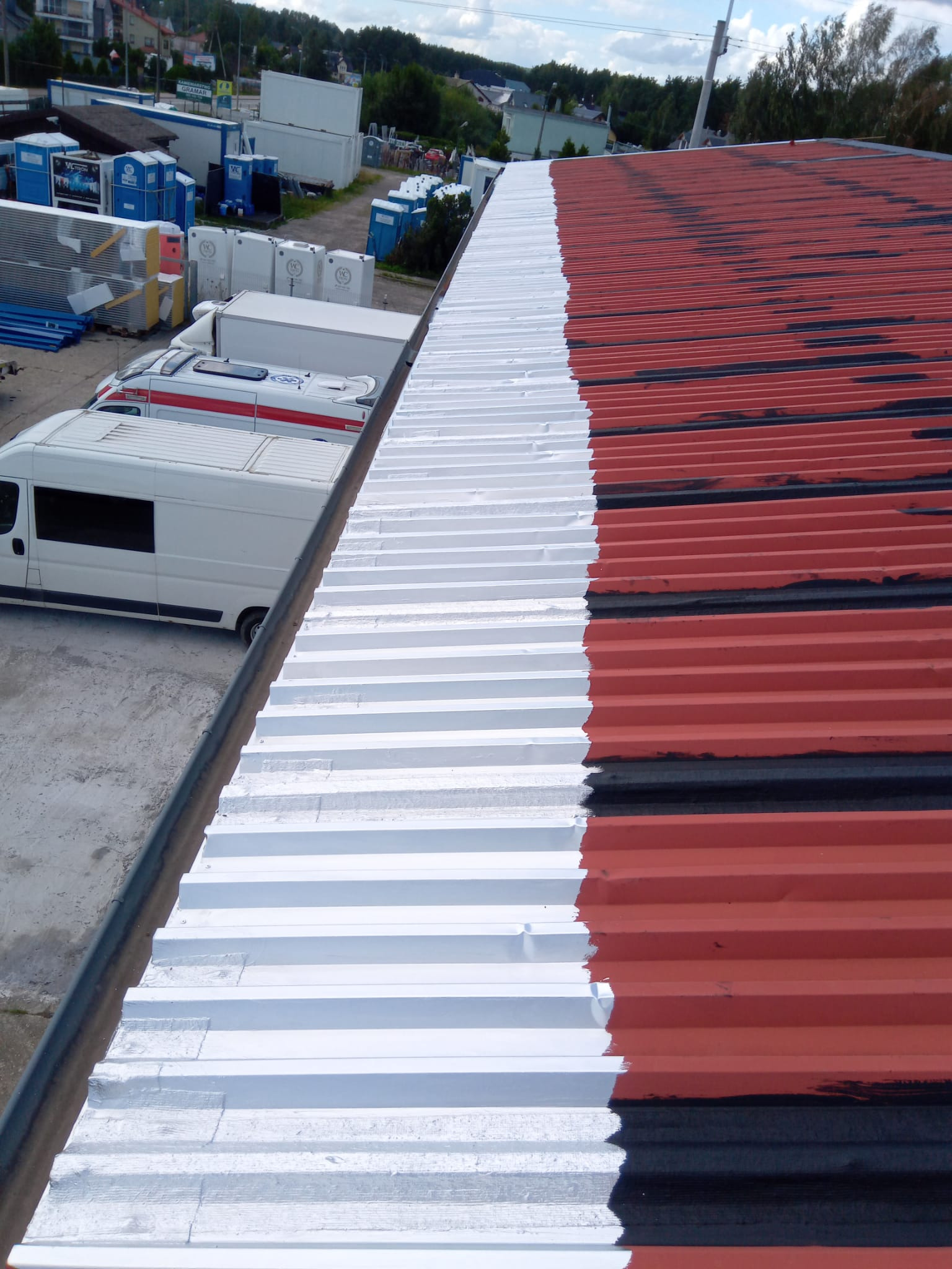 Częściowo pomalowany blaszany dach na biało, z widocznymi fragmentami czerwieni i czerni. W tle zaparkowane busy i kontenery.