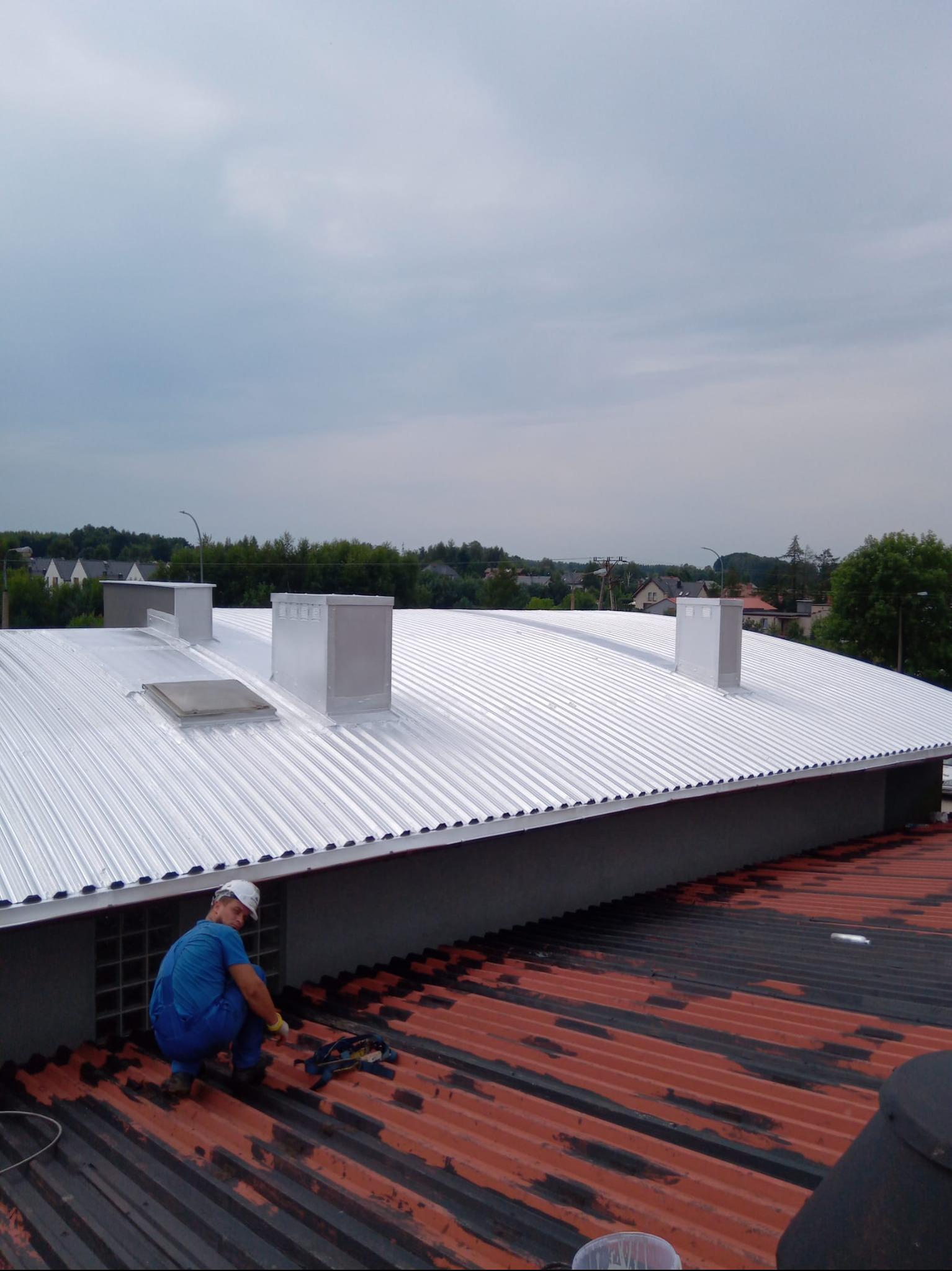 Dekarz w trakcie pracy na dachu z blachy trapezowej, widoczne kominy wentylacyjne i okno dachowe. Stary dach z blachy trapezowej w kolorze czerwono-czarnym.