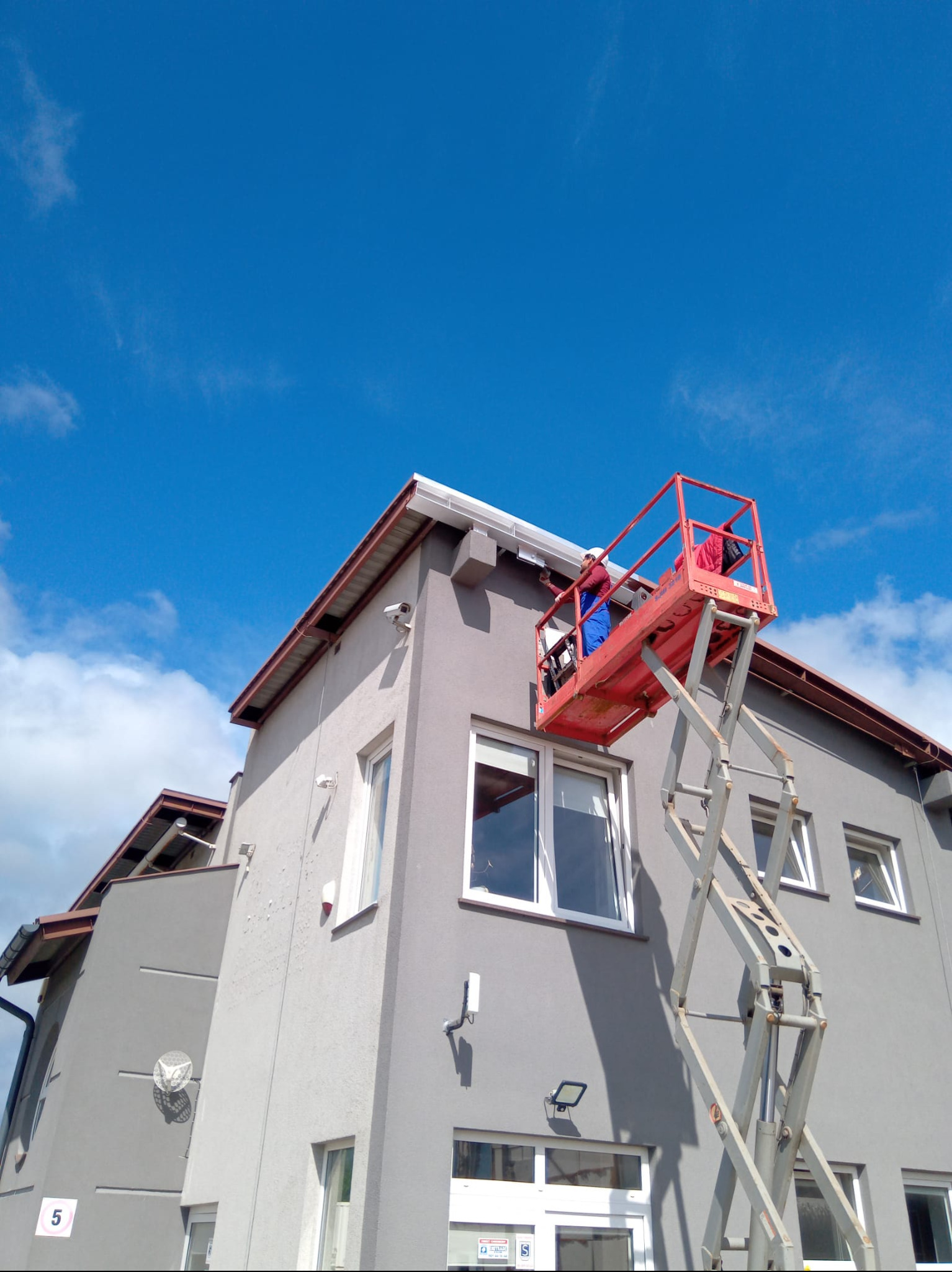 Pracownik na podnośniku koszowym montuje orynnowanie na szarym budynku z brązowym dachem na tle błękitnego nieba z chmurami. Widok z dołu.