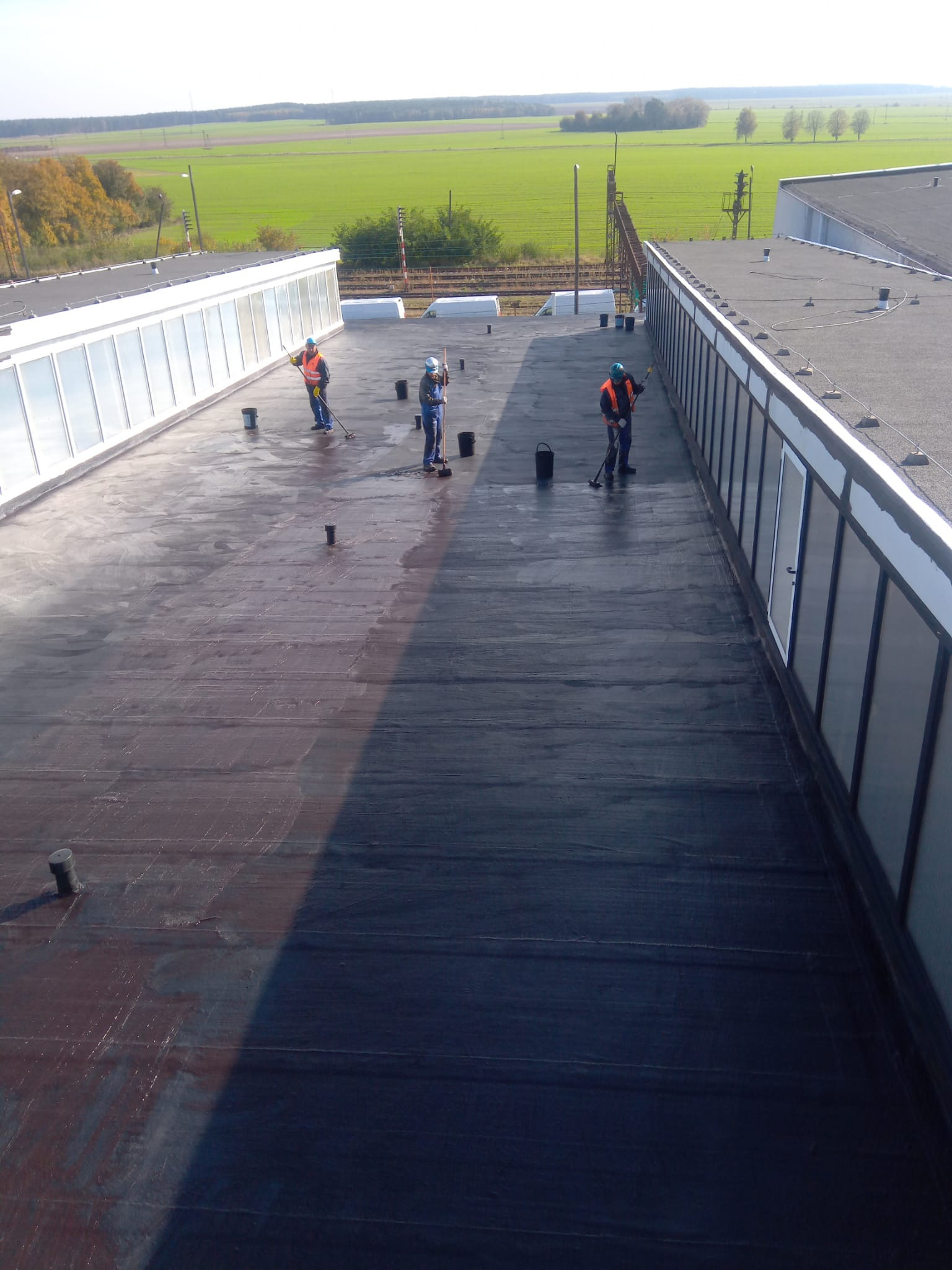 Praca na dachu budynku przemysłowego: trzech dekarzy w odblaskowych kamizelkach rozprowadza masę bitumiczną na płaskiej powierzchni dachu. Widok z góry, w tle zielone pola.