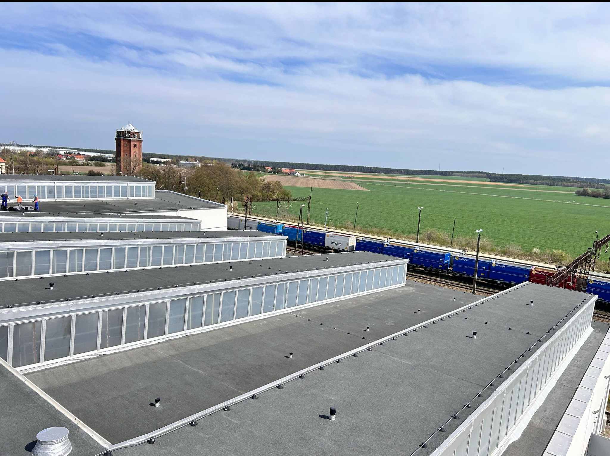 Rozległy, płaski dach przemysłowy z lukarnami, widok z góry na tle zielonych pól i błękitnego nieba. Na dachu widoczni pracownicy w trakcie pracy. W tle wieża ciśnień.