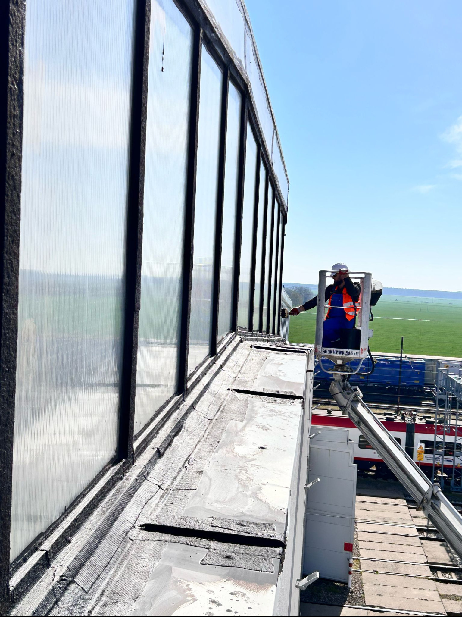 Pracownik w podnośniku koszowym naprawia dach z paneli na tle zielonych pól i błękitnego nieba. Widoczne narzędzia i fragment pociągu.