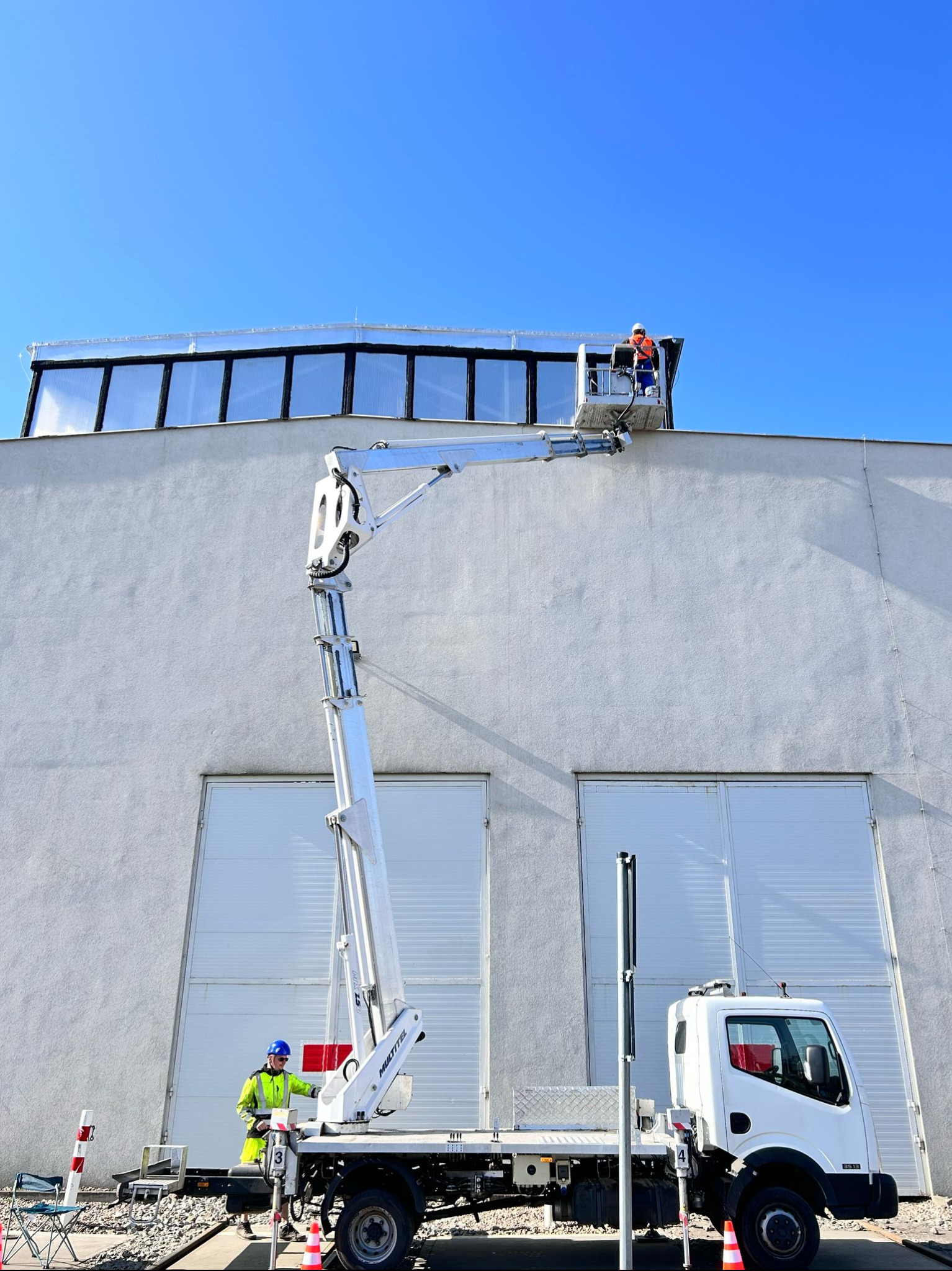 Pracownik na podnośniku koszowym montuje okna dachowe na hali przemysłowej. Widoczny biały budynek z szarym dachem i błękitne niebo.