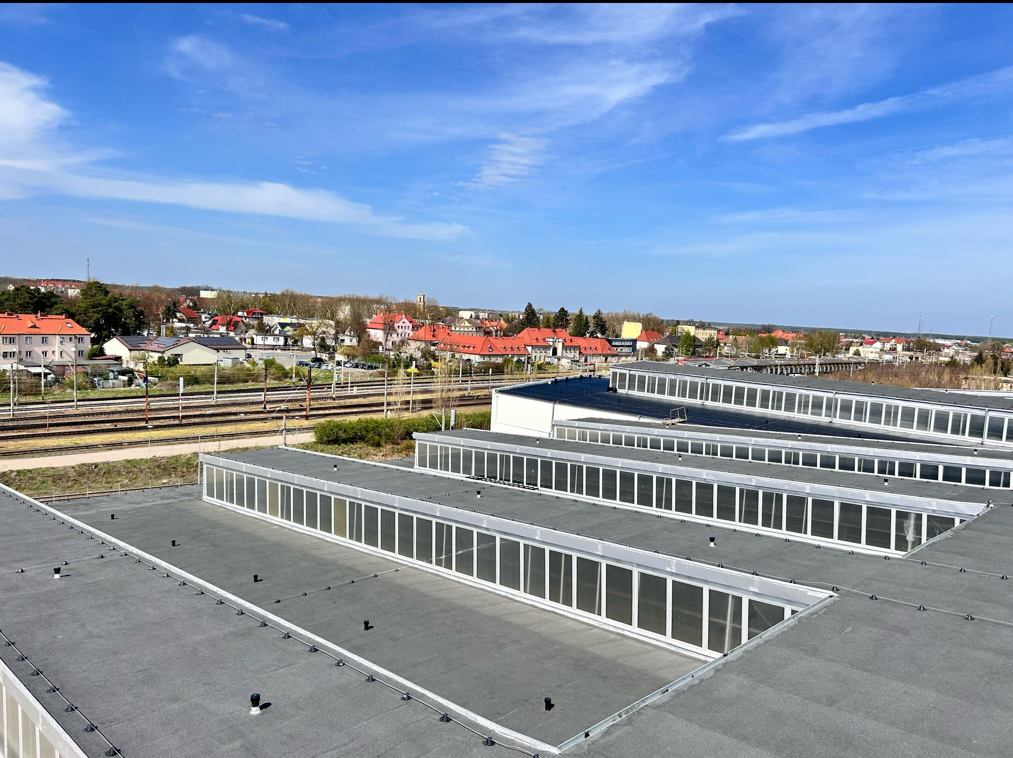 Płaski dach budynku przemysłowego z świetlikami, widok z góry na tle zabudowy miejskiej i błękitnego nieba. Dach pokryty papą, z widocznymi elementami mocującymi.