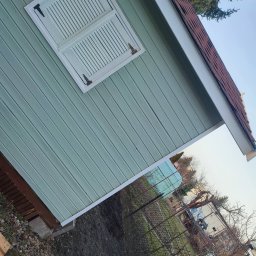 Mateusz Nowak - Ściana pomalowana na pastelowy zielony kolor z białymi okiennicami i brązowym dachem. Widoczne drewniane panele i elementy wykończeniowe.