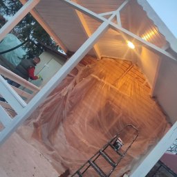 Mateusz Nowak - Malowanie altany na biało. Widoczny malarz w trakcie pracy, drabina i folia ochronna wewnątrz konstrukcji. Ujęcie z dołu, podkreślające geometrię altany.