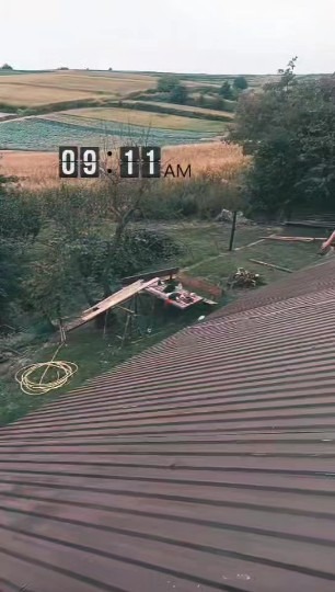 Widok z góry na brązowy dach z blachy falistej, z widocznym rusztowaniem i narzędziami w tle, oraz polami uprawnymi w oddali. Godzina 09:11 AM.