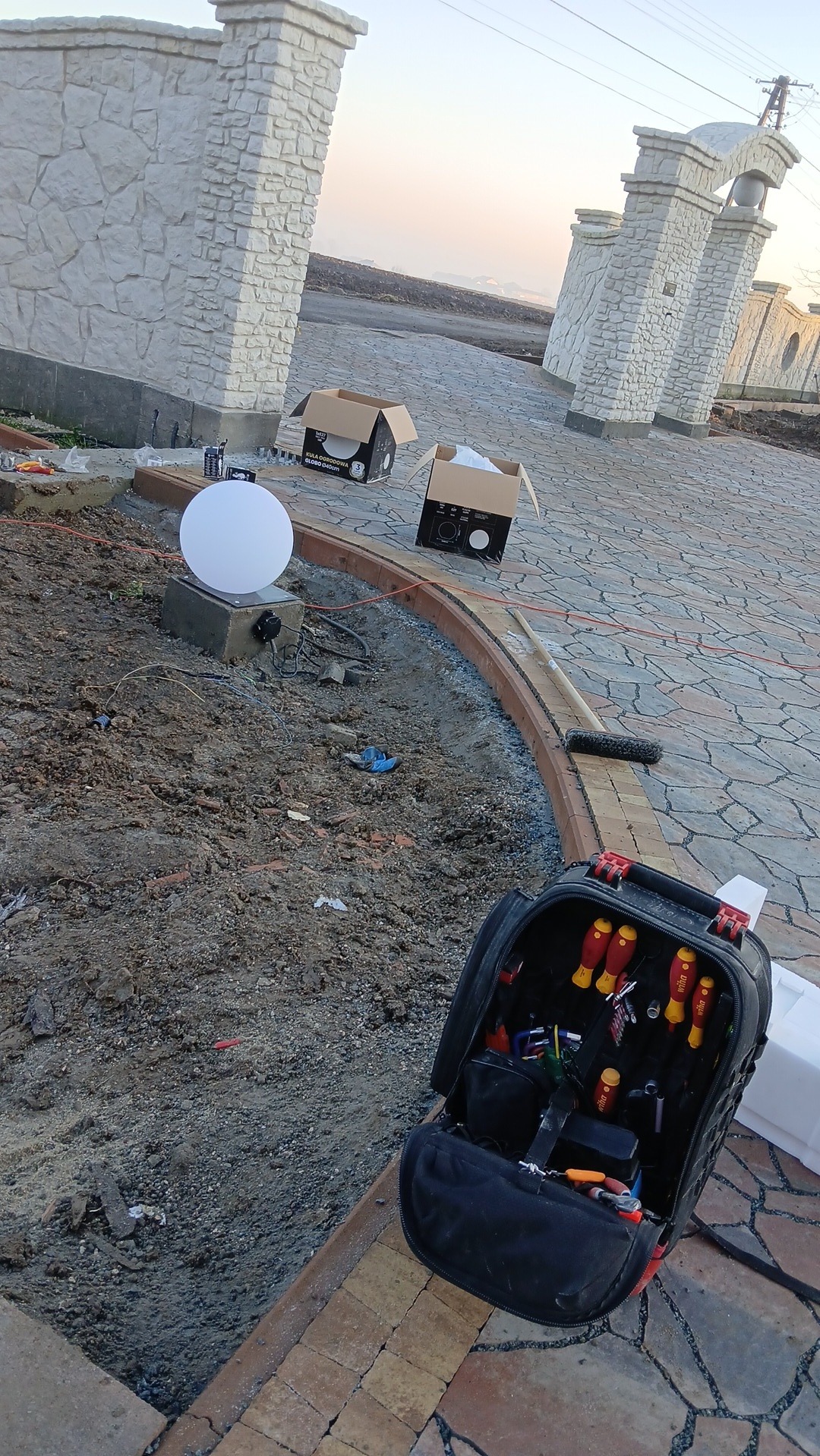 Montaż oświetlenia ogrodowego: kula świetlna na betonowym postumencie, narzędzia w torbie, przygotowane kable i otwarte kartony na tle kamiennej ścieżki i ogrodzenia.