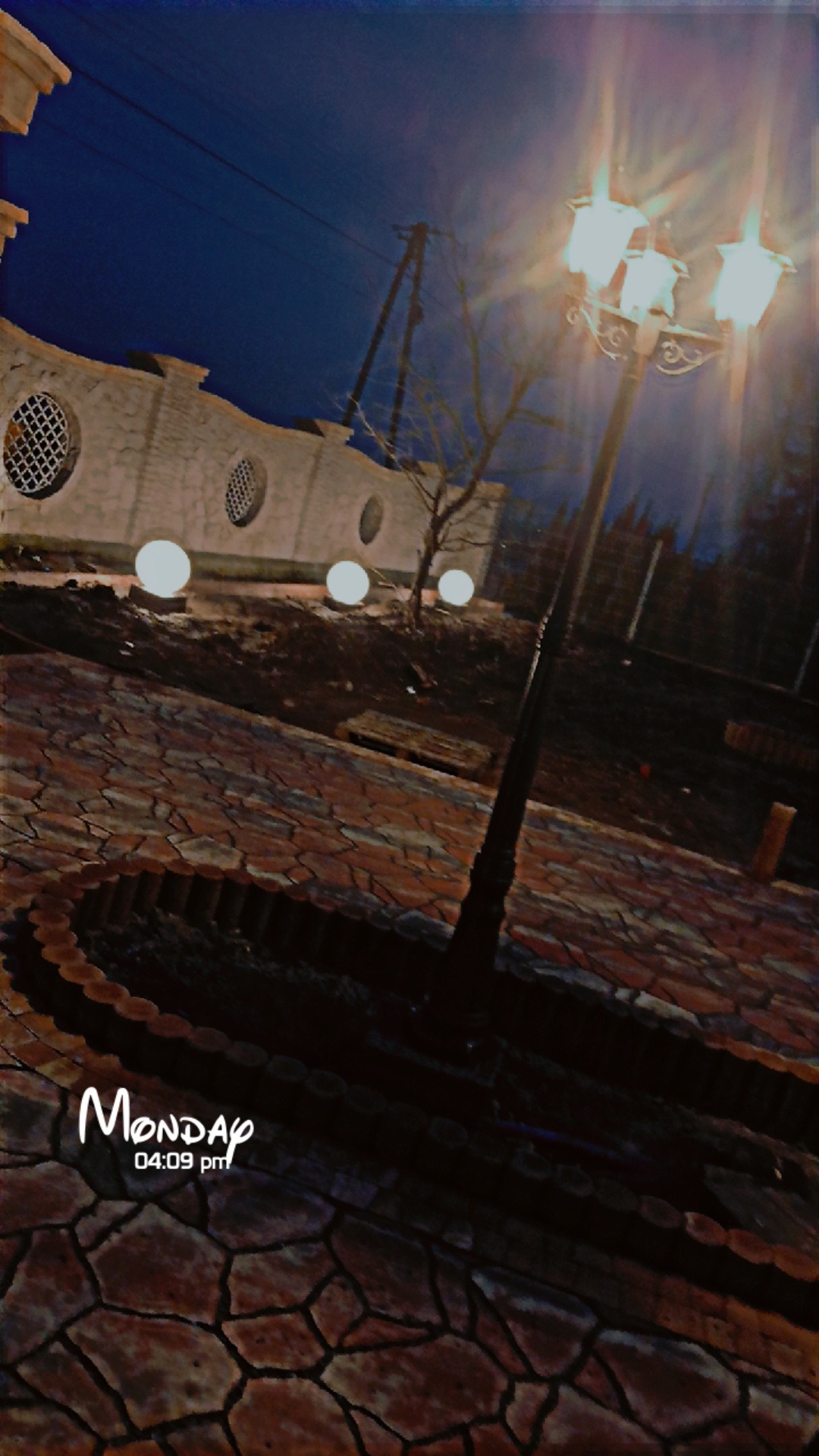 Oświetlony park nocą: brukowa ścieżka, lampa uliczna z trzema kloszami, budynek z okrągłymi oknami i kuliste lampy. Ujęcie z dołu, widoczny fragment daty.