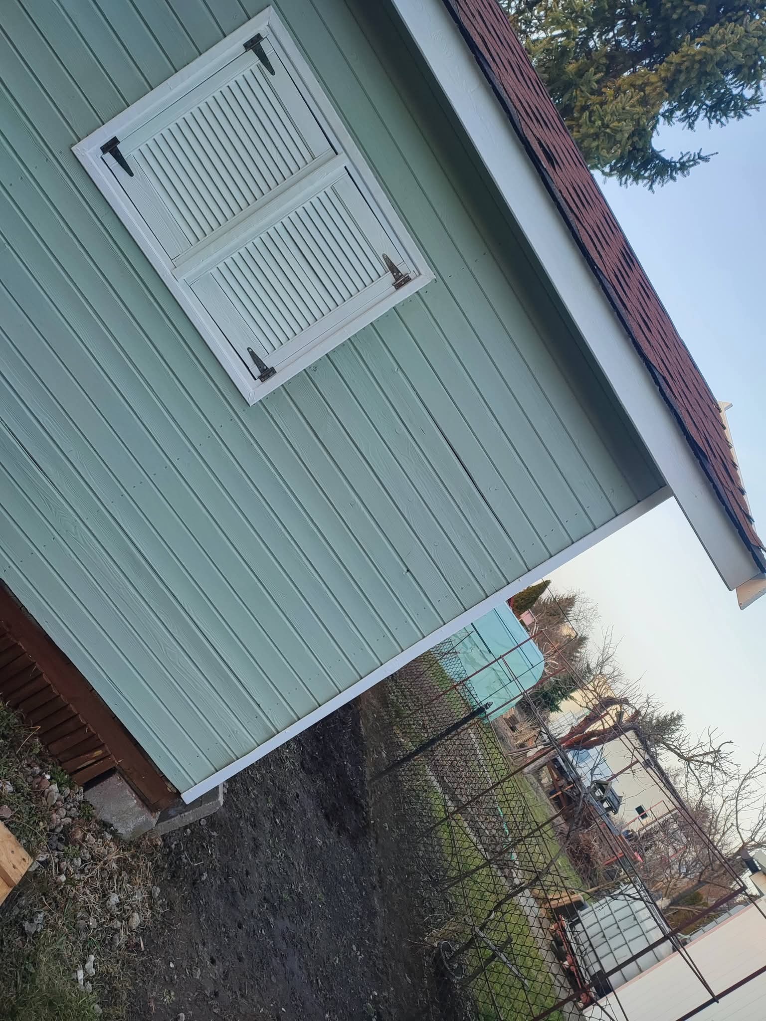 Ściana pomalowana na pastelowy zielony kolor z białymi okiennicami i brązowym dachem. Widoczne drewniane panele i elementy wykończeniowe.