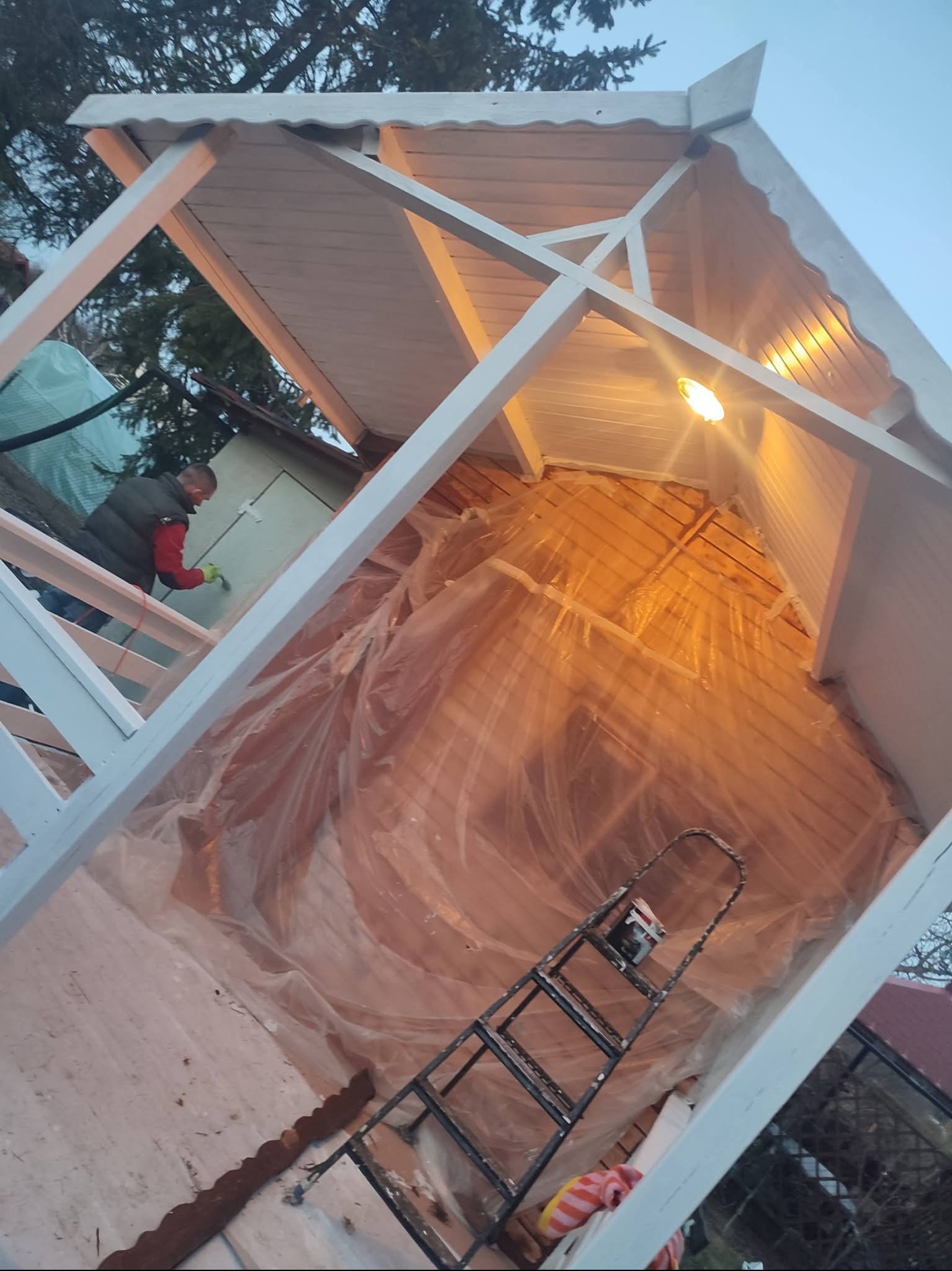 Malowanie altany na biało. Widoczny malarz w trakcie pracy, drabina i folia ochronna wewnątrz konstrukcji. Ujęcie z dołu, podkreślające geometrię altany.