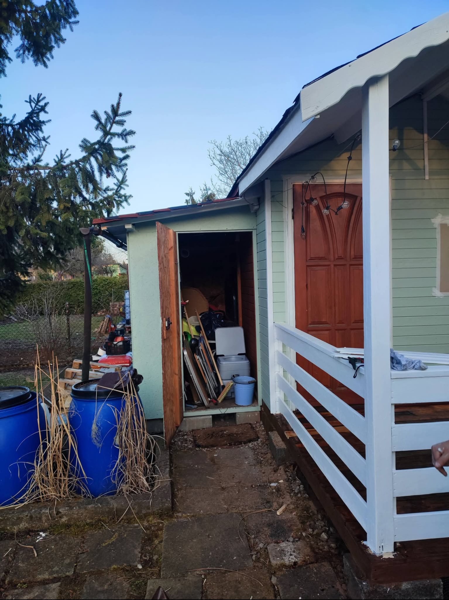 Otwarty, zagracony domek narzędziowy z niebieskimi beczkami na deszczówkę i świeżo pomalowaną werandą w ogrodzie. Widoczne narzędzia i akcesoria ogrodowe.