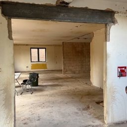 Wyburzenia Mazowieckie Mateusz Zalewski - Wnętrze po usunięciu ściany działowej, widoczna stalowa belka wzmacniająca, surowe ściany i podłoga, okno z czarną ramą w tle, krzesła i pojemnik w rogu pomieszczenia.