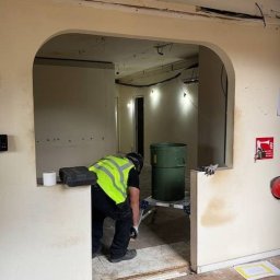 Wyburzenia Mazowieckie Mateusz Zalewski - Pracownik w kamizelce przy wyburzonym łuku ściany, widoczne narzędzia i pojemnik na odpady. Ślady po usuniętej ścianie, surowy stan wnętrza.