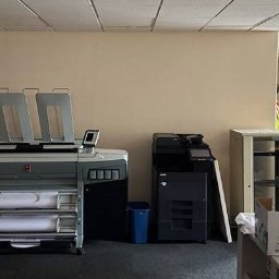 Wyburzenia Mazowieckie Mateusz Zalewski - Wnętrze biura z dużym ploterem, drukarką i szafką. Widoczny kosz na śmieci i kartony. Ściany w kolorze beżowym, sufit biały z widocznymi kablami.