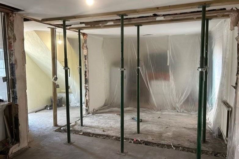 Wnętrze w trakcie prac rozbiórkowych z podporami stropowymi, zabezpieczone folią. Widoczne ślady po usuniętych ścianach i pozostałości gruzu na podłodze.