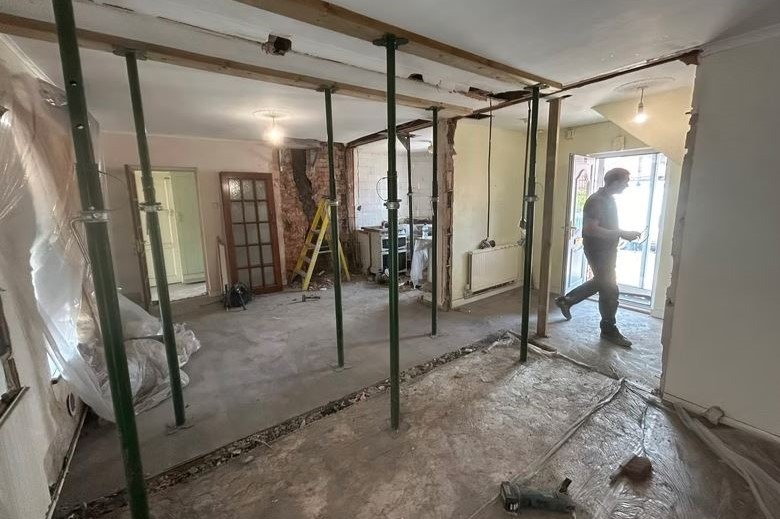 Wnętrze w trakcie wyburzania ścian, podparte stemple budowlane, widoczny gruz i narzędzia. Mężczyzna w tle przechodzi przez otwór drzwiowy.