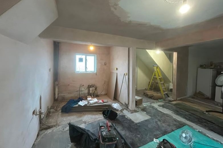 Wnętrze w trakcie remontu: otynkowane ściany, okno, instalacje, narzędzia, drabina i materiały budowlane. Widoczne ślady prac przygotowawczych do dalszych etapów.