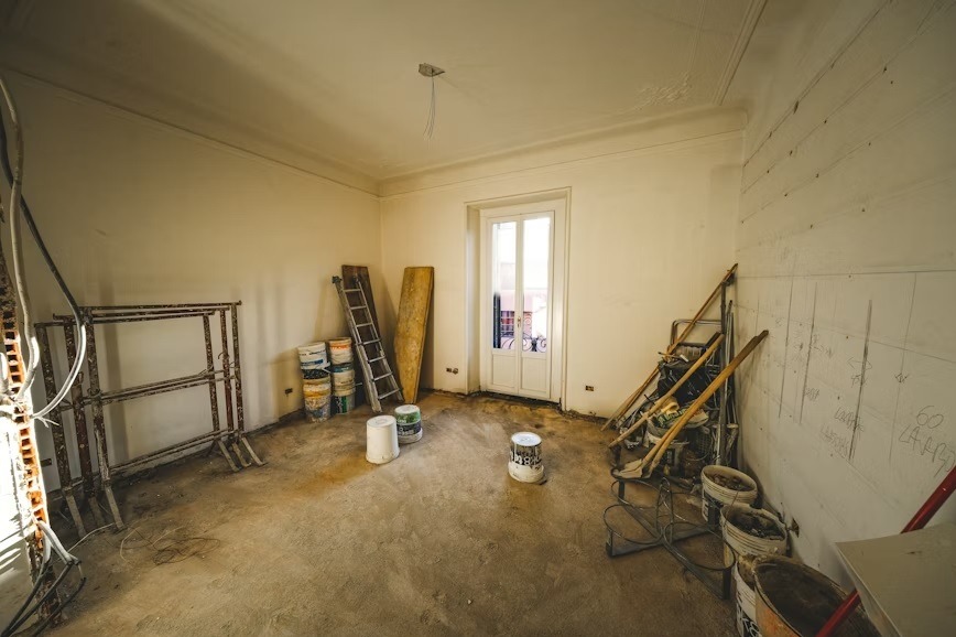 Wnętrze pokoju w trakcie remontu: widoczne rusztowania, drabina, wiadra z farbą i ślady po instalacjach na ścianach. Przygotowanie do prac wykończeniowych.