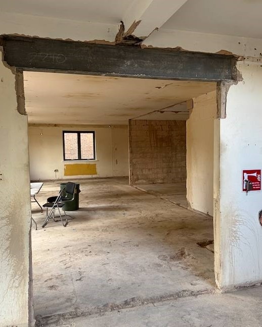 Wnętrze po usunięciu ściany działowej, widoczna stalowa belka wzmacniająca, surowe ściany i podłoga, okno z czarną ramą w tle, krzesła i pojemnik w rogu pomieszczenia.