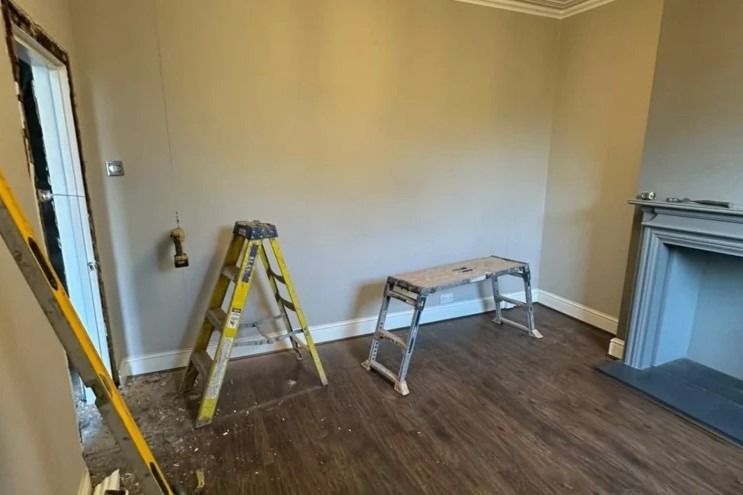 Przygotowania do prac remontowych w Warszawie: drabina, poziomica przy ościeżnicy, wisząca wiertarka. Pokój z drewnianą podłogą i kominkiem.