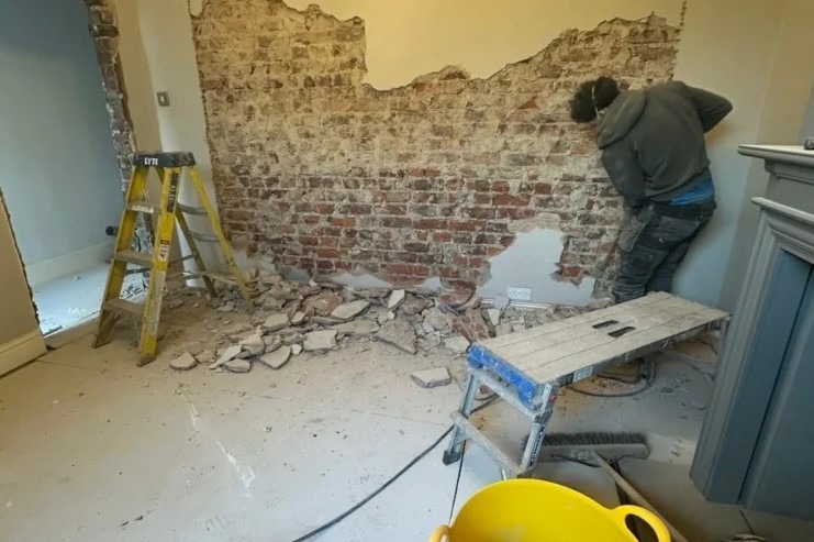 Ceglana ściana w trakcie demontażu, widoczne narzędzia i gruz. Pracownik w trakcie pracy. Żółty kubeł na gruz w rogu kadru.