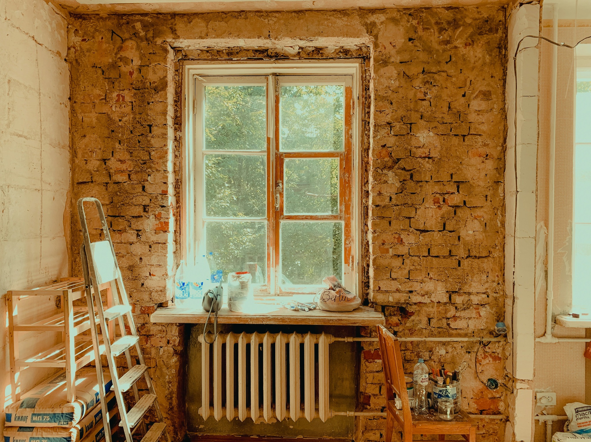 Ceglana ściana w trakcie renowacji z oknem, drabina oparta o ścianę, worki z zaprawą, narzędzia na parapecie i krześle. Widoczne ślady po skuciu tynku.