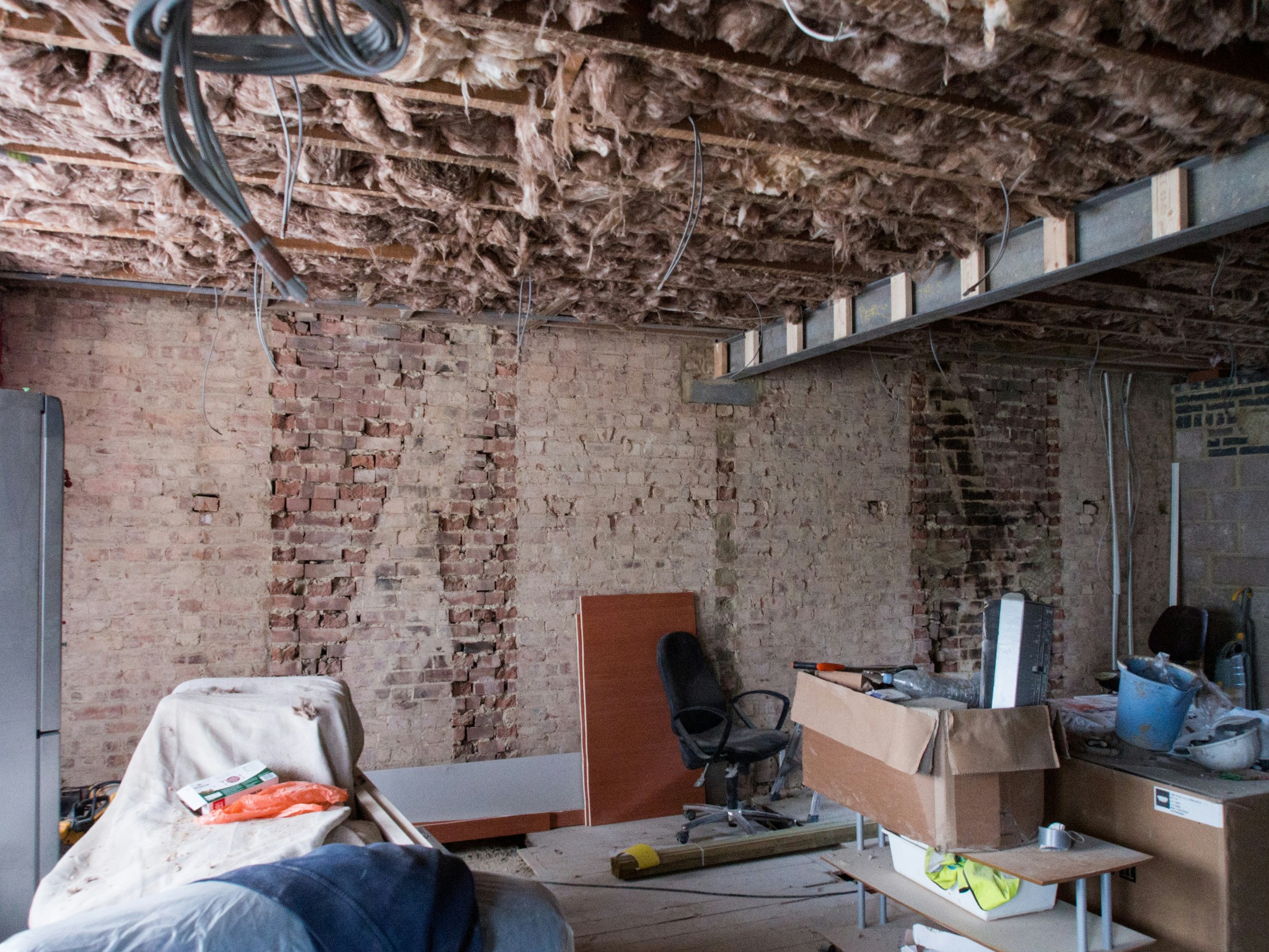 Odsłonięta ceglana ściana i sufit z izolacją z waty szklanej w trakcie prac remontowych. Widoczne kable, meble i narzędzia w pomieszczeniu.