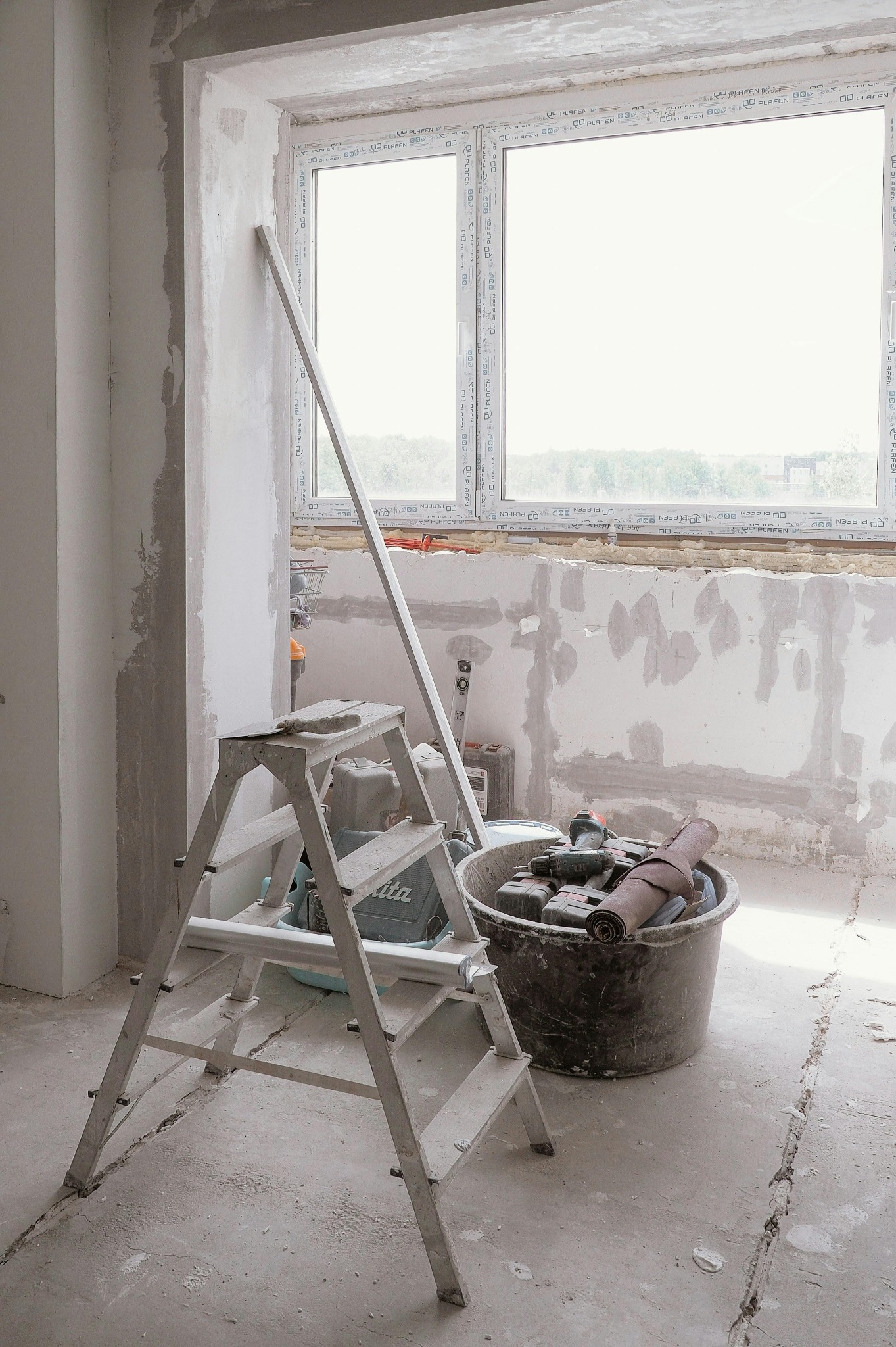 Wnętrze w trakcie remontu: drabina, wiadro z narzędziami, ściana z widocznymi śladami prac, okno z widokiem na zewnątrz. Przygotowanie do dalszych prac wykończeniowych.