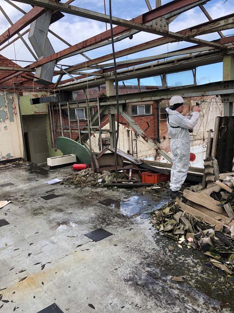 Wnętrze budynku po rozbiórce z widocznymi elementami konstrukcyjnymi i gruzem. Osoba w kombinezonie ochronnym dokumentuje postęp prac. Widoczne pozostałości ścian i sufitu.