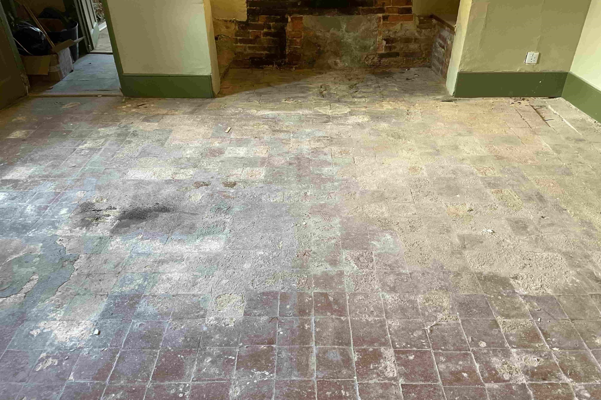 Pokój po demontażu podłogi z odsłoniętą, starą, kwadratową posadzką. Widoczne ślady po usuniętych elementach i fragmenty ceglanej ściany w tle.